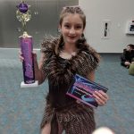A girl in a costume holding a trophy at Carlie's Art Gallery.