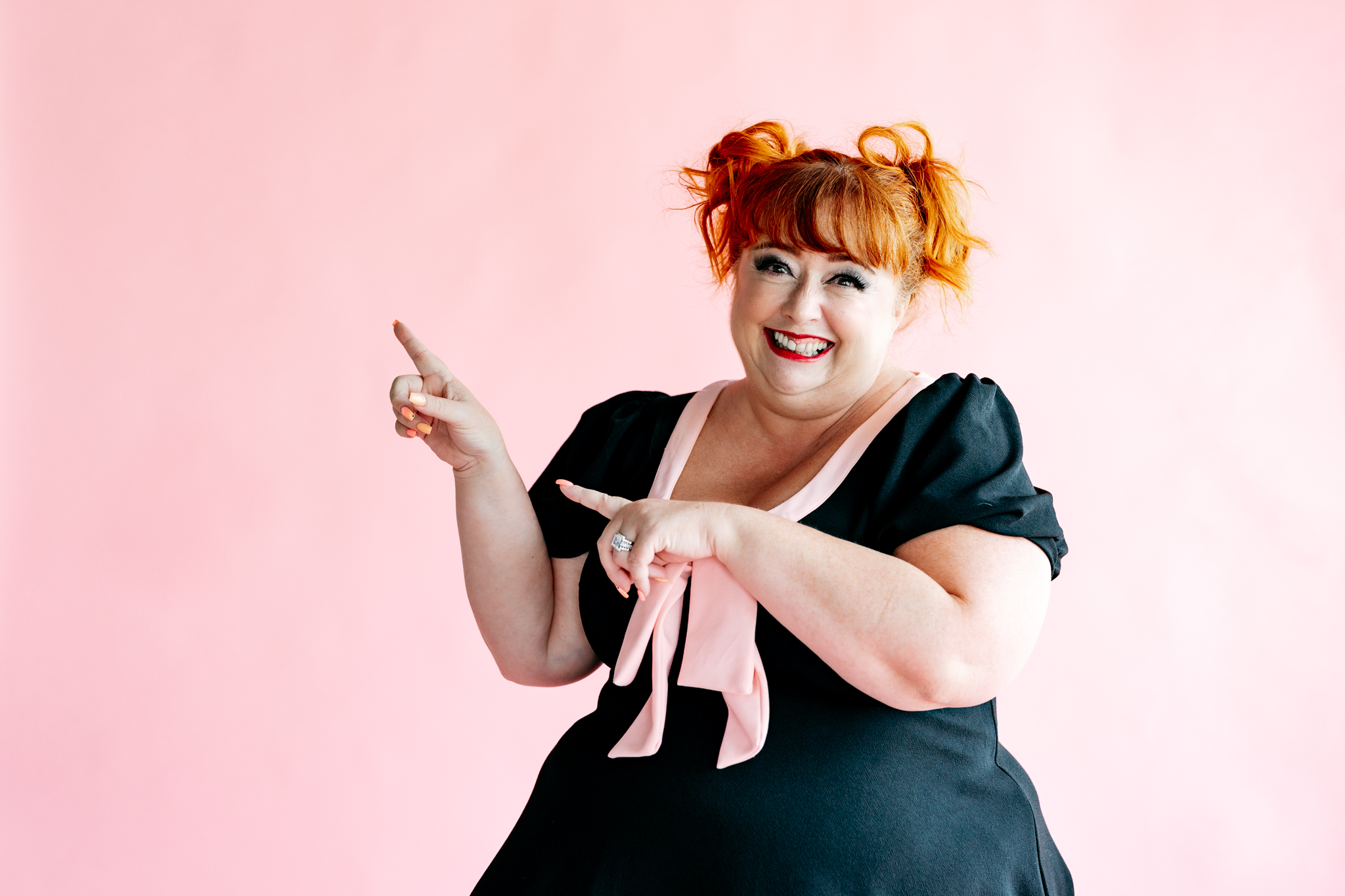 Virtual Work Wife - A person with red hair in playful pigtails smiles and points to the right while wearing a black dress with a pink ribbon, standing against a pink background, symbolizing the spirit of Discovery 2.0 and innovation in a vibrant world.