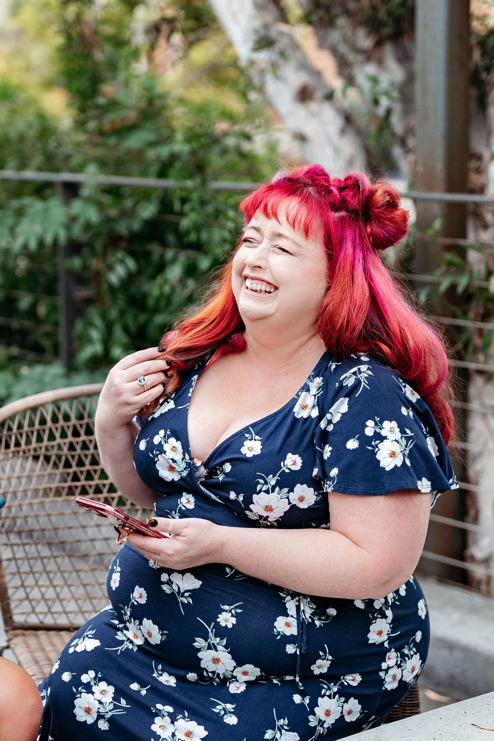 Virtual Work Wife - A joyful woman with red hair styled in buns, wearing a floral dress, laughing while sitting on a patio chair, perfectly illustrating an efficient tagging system for online content categorization.