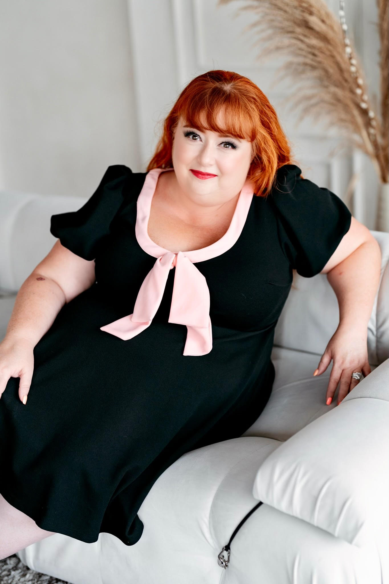 Virtual Work Wife - A woman with red hair, wearing a black dress with a light pink bow, is seated on a white couch, looking every bit the ideal virtual work wife.