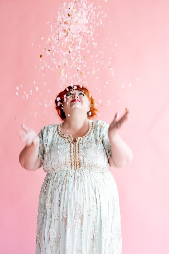 Virtual Work Wife - A woman with short red hair in a light, embroidered dress throws confetti into the air against a pink background, embodying the joy of having a Virtual Work Wife to share successes and celebrations with.