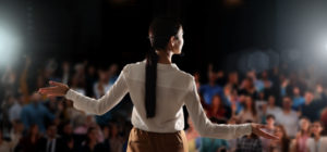 Virtual Work Wife - A person with a headset gestures while speaking to a blurred audience under bright lights.